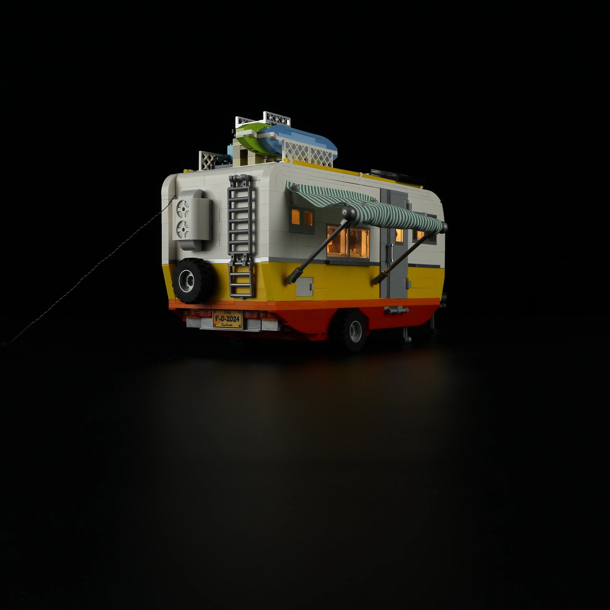 Lumibricks F9032 Travel Trailer rear view at night with illuminated windows, striped awning, rooftop surfboards, and ladder
